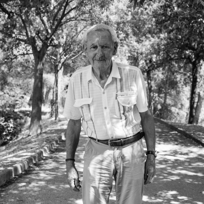 Un homme âgé marche tranquillement dans un parc, entouré d'arbres et de feuillage. Il porte une chemise légère à manches courtes et un pantalon clair, tenant une posture droite et confiante. La lumière du soleil filtre à travers les branches, créant des motifs d'ombres sur le sol.